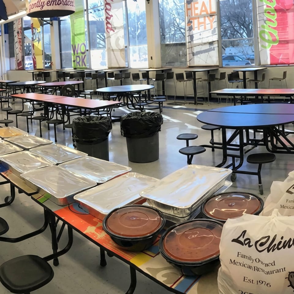 School cafeteria with empty tables and chairs, featuring covered catering trays, bowls of salsa, and La Chinita Mexican restaurant bags.