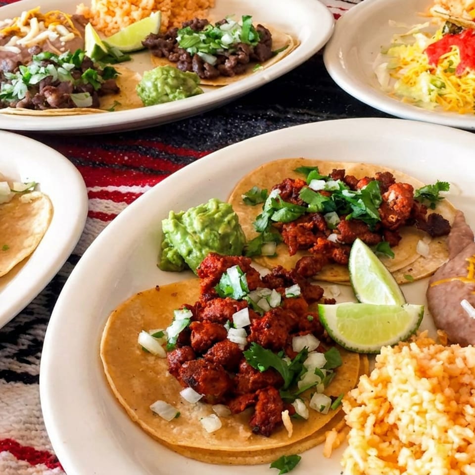 Assorted Mexican dishes featuring tacos, rice, beans, guacamole, lime wedges, and a Modelo beer on a patterned tablecloth.