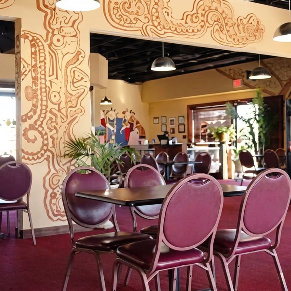 Spacious retro restaurant dining room with burgundy chairs, dark tables, patterned murals, pendant lights, and a service counter.