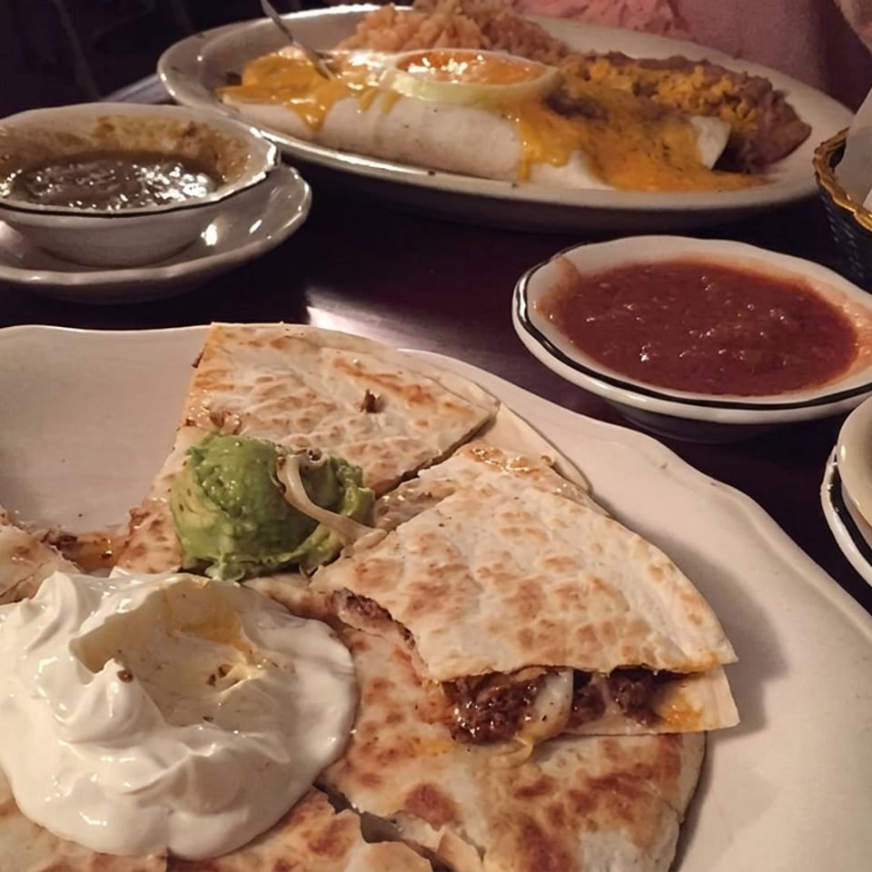 Mexican restaurant table featuring quesadillas with guacamole and sour cream, enchiladas, beans, tortilla chips, salsa, and a drink.